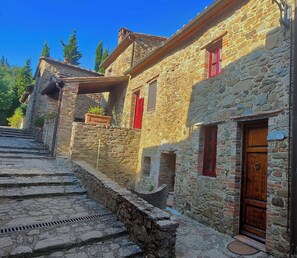 A stone building with a wooden door and red shutters, featuring a stone staircase leading up to the entrance.