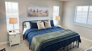 Une chambre à coucher avec un grand lit, deux tables de chevet, une commode, un tableau au mur et des fenêtres équipées de stores.