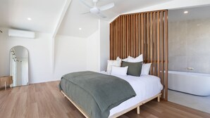 Un dormitori modern amb llit, paret de lamel·les de fusta, un mirall gran, ventilador de sostre i banyera.