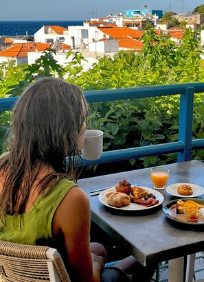 Una persona seduta a tavola con prodotti per la colazione e un bicchiere di succo d'arancia, con vista su una cittadina costiera caratterizzata da edifici con tetti rossi.