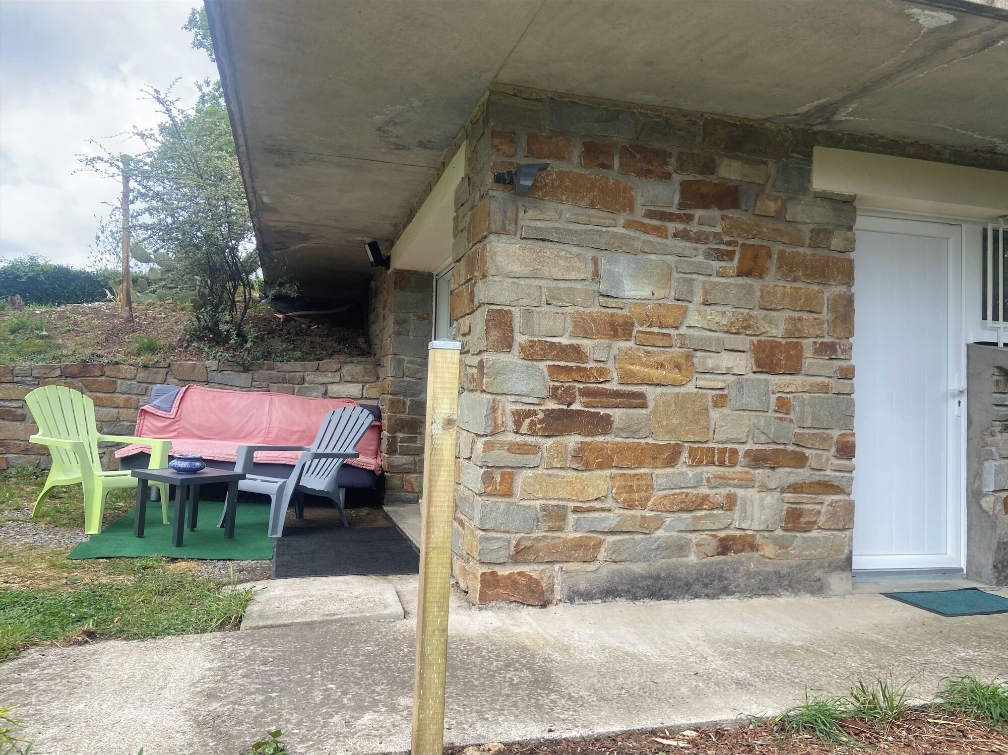 A stone exterior wall with a door, a covered outdoor seating area with plastic chairs and a table, and a concrete pathway leading to the door.