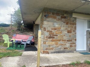 A stone exterior wall with a door, a covered outdoor seating area with plastic chairs and a table, and a concrete pathway leading to the door. - Private studio in the countryside, located in a local resident's home (Cordemais)