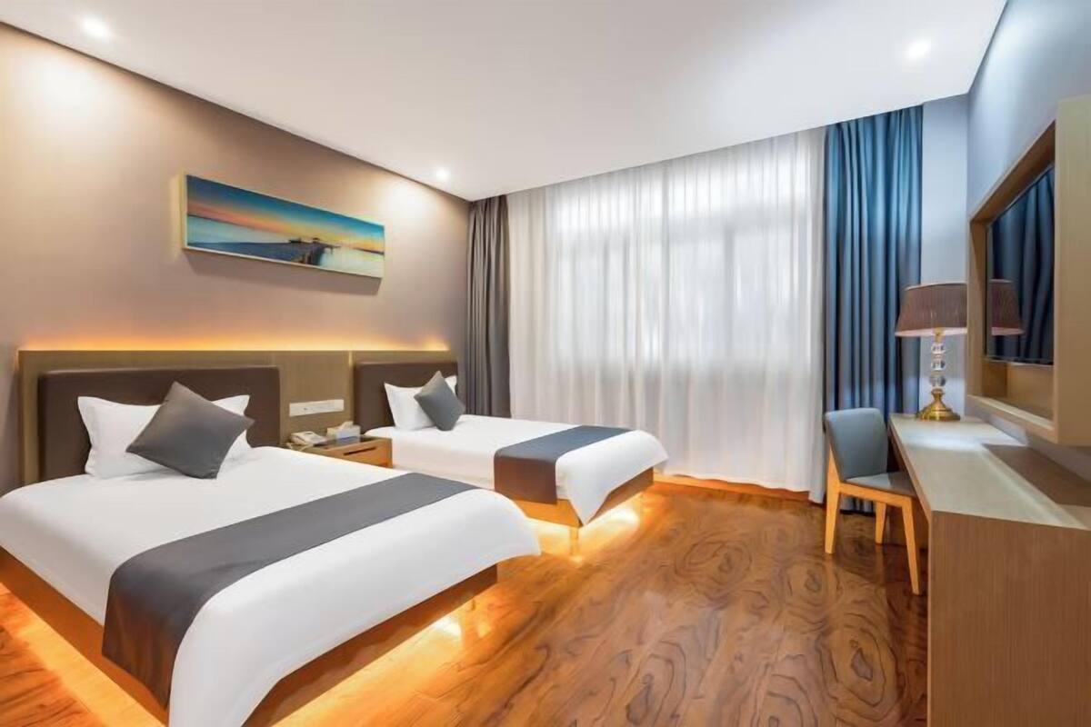A hotel room with two beds, a desk, a chair, a lamp, a painting on the wall, and a large window with curtains.
