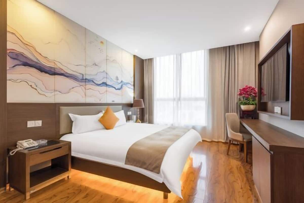 A hotel room with a bed, bedside table, lamp, chair, and a large abstract painting on the wall.