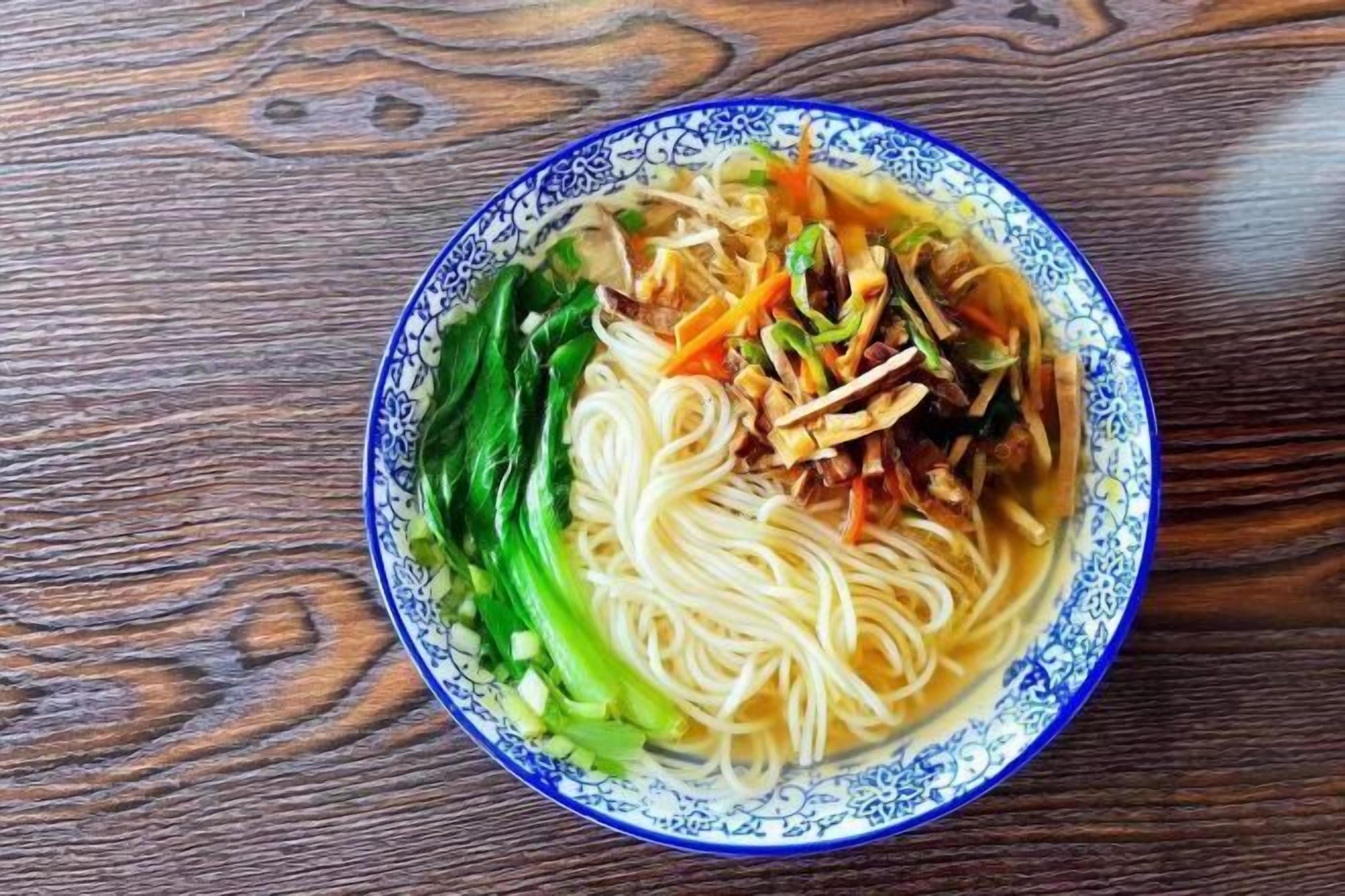 A bowl of noodles with vegetables on a wooden table.