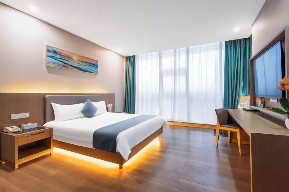 A hotel room with a bed, bedside table, desk, chair, and a large window with curtains.