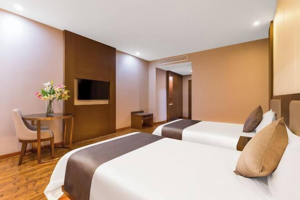 A hotel room with two beds, a small table with a vase of flowers, a TV mounted on the wall, and a wooden floor.