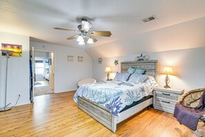Une chambre avec un plancher en bois, un lit recouvert d’une couvre-lit à motifs bleus et blancs, deux lampes de chevet, un ventilateur de plafond et une table de chevet avec une lampe.