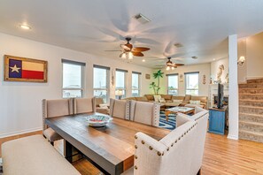 Un salon spacieux avec une grande table à manger en bois, des chaises recouvertes d’un tissu beige et un ventilateur de plafond.
