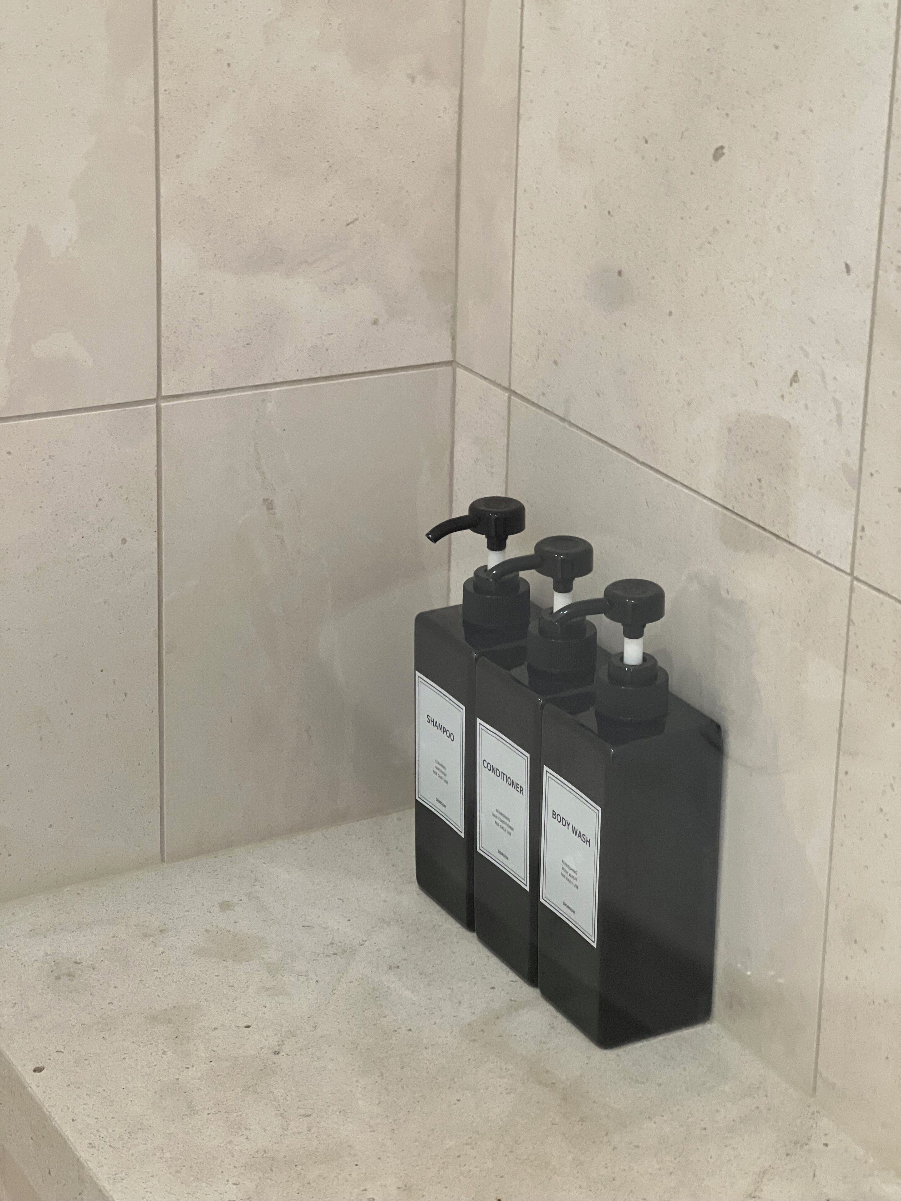 Casa de banho com parede revestida em azulejos e um prateleira com três frascos pretos rotulados como champô, condicionador e gel de banho.