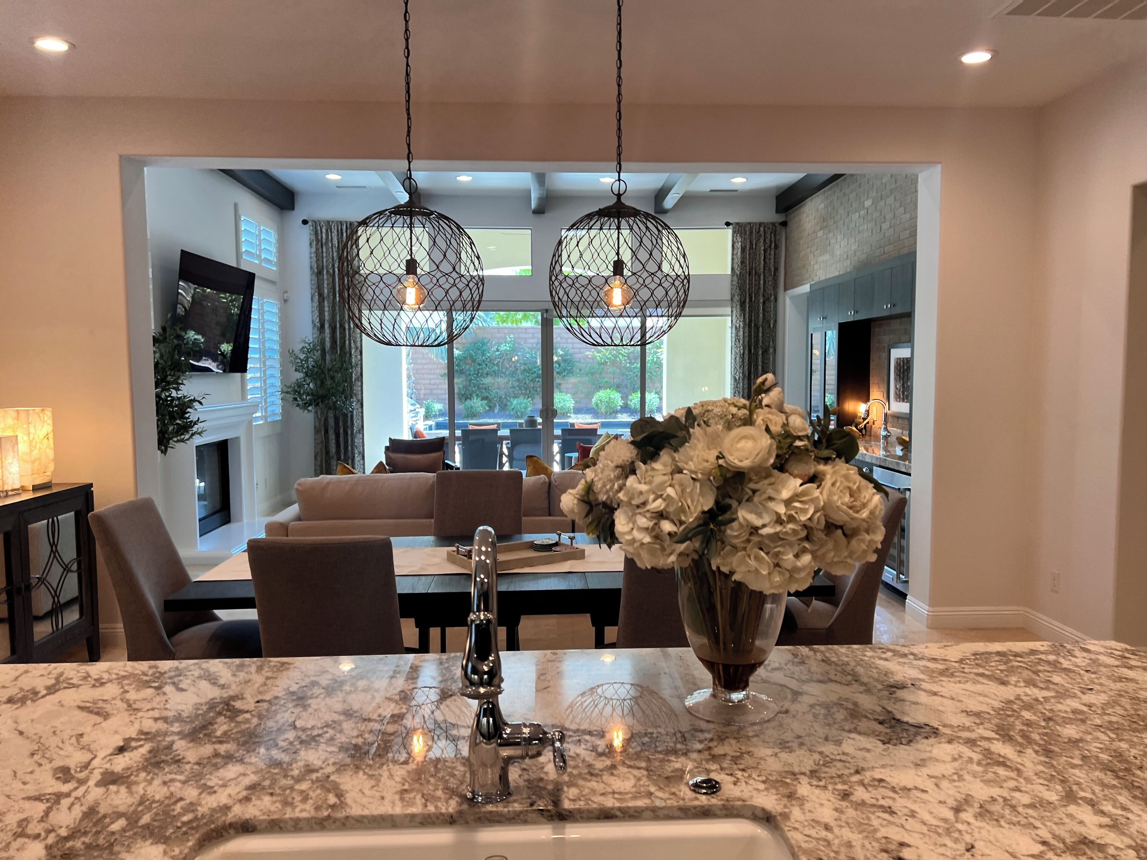 A modern kitchen with a marble countertop, a sink, and a vase of flowers. There is a dining area with a sofa and chairs visible through an open archway.