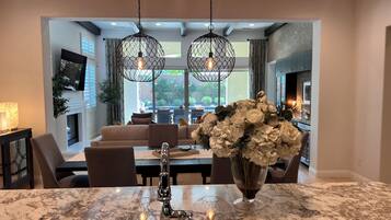 A modern kitchen with a marble countertop, a sink, and a vase of flowers. There is a dining area with a sofa and chairs visible through an open archway.