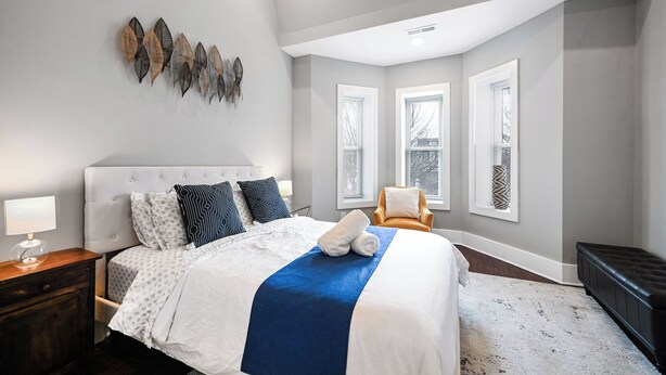A modern bedroom with a large bed, a nightstand, a chair, and a window with blinds. - Bright Washington DC Apartment (Washington)