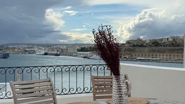 Eine Terrasse mit einem runden Tisch, zwei Stühlen, einer Ziervase und Blick auf eine Stadtlandschaft und Wasser.