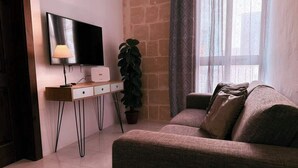 Ein Wohnzimmer mit einer Couch, einem Fernseher, einem Schreibtisch mit Lampe und einer Topfpflanze.
