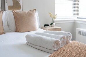Een net opgemaakt bed met opgerolde witte handdoeken, een nachtkastje met een klok, en een raam met luxaflex.