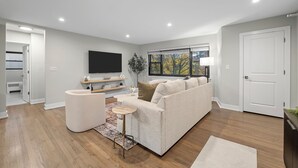 Una sala de estar moderna con un sofá, una mesa de centro y un televisor. Se ve una ventana con vistas a árboles y una puerta a la izquierda.