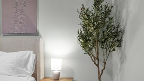 Una cama bien tendida con sábanas blancas, una mesita de noche con una lámpara y una planta alta en maceta junto a una pared.