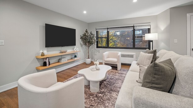 Una sala de estar moderna con un televisor de pantalla plana, sofás blancos, una mesa de centro y un amplio ventanal con vistas a árboles.
