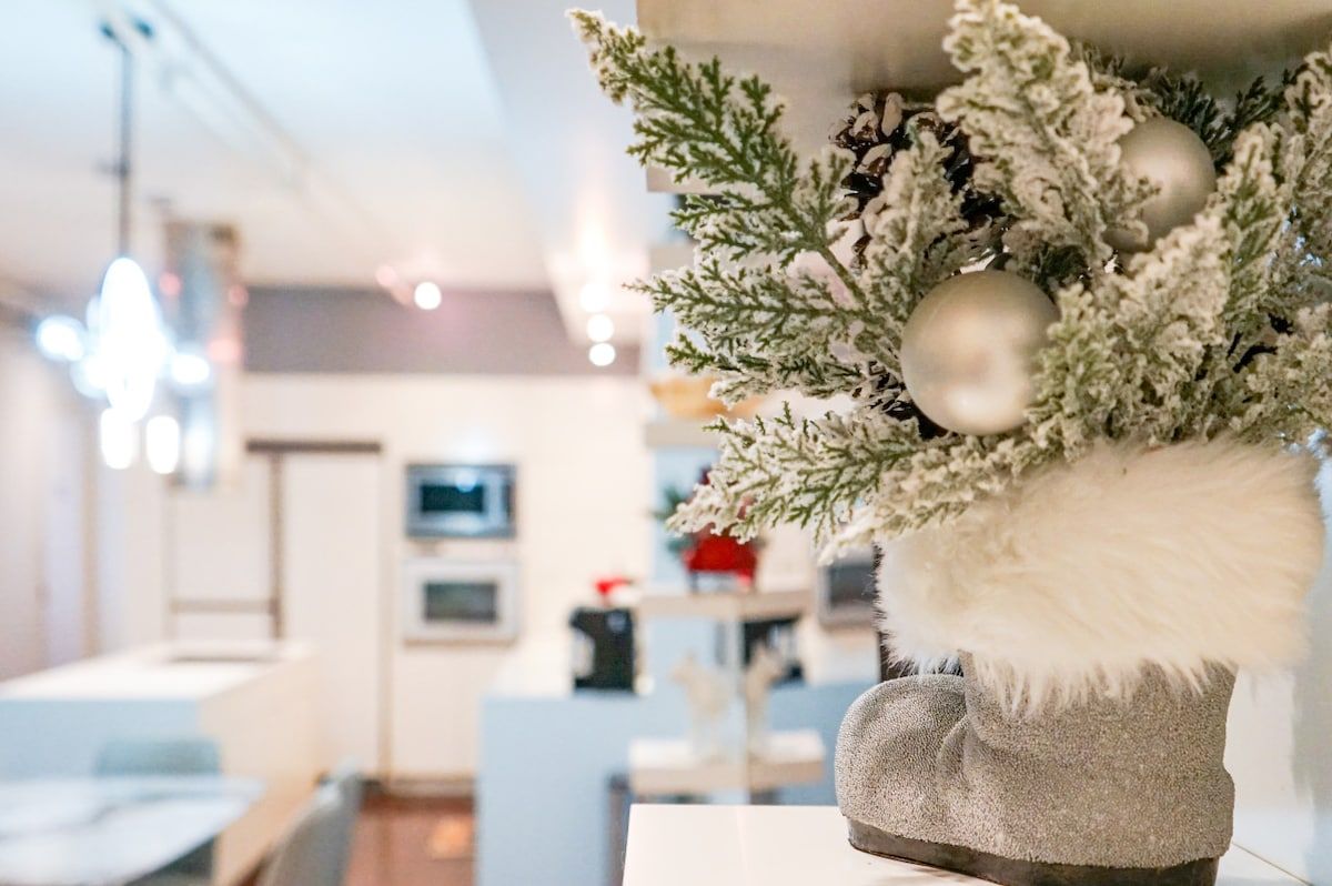 Un interior moderno con una planta decorativa en una maceta texturizada, una zona de comedor con una mesa blanca y una cocina al fondo.