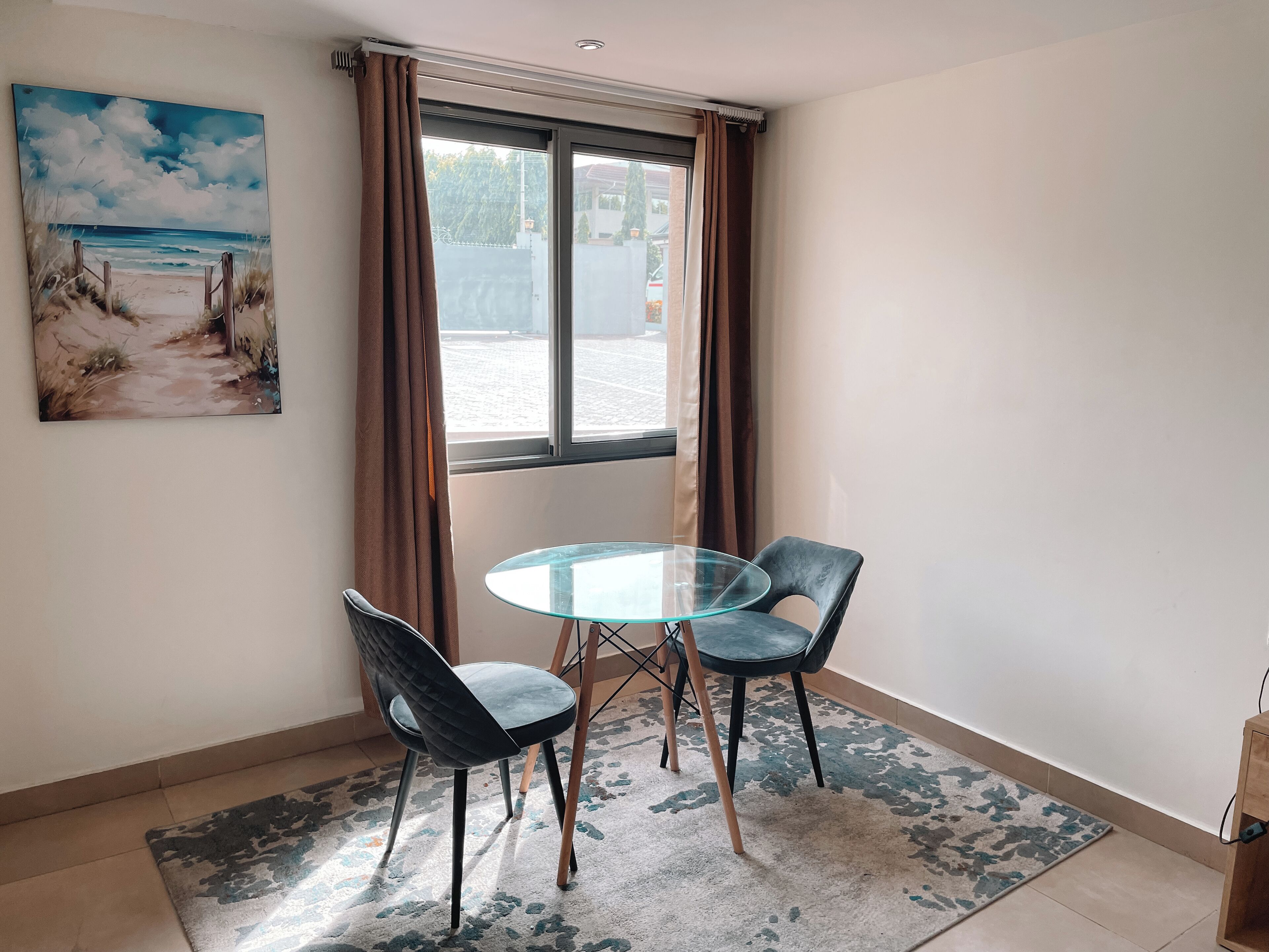 A room with a glass table and two chairs, a painting of a beach scene, and a view of the street outside.
