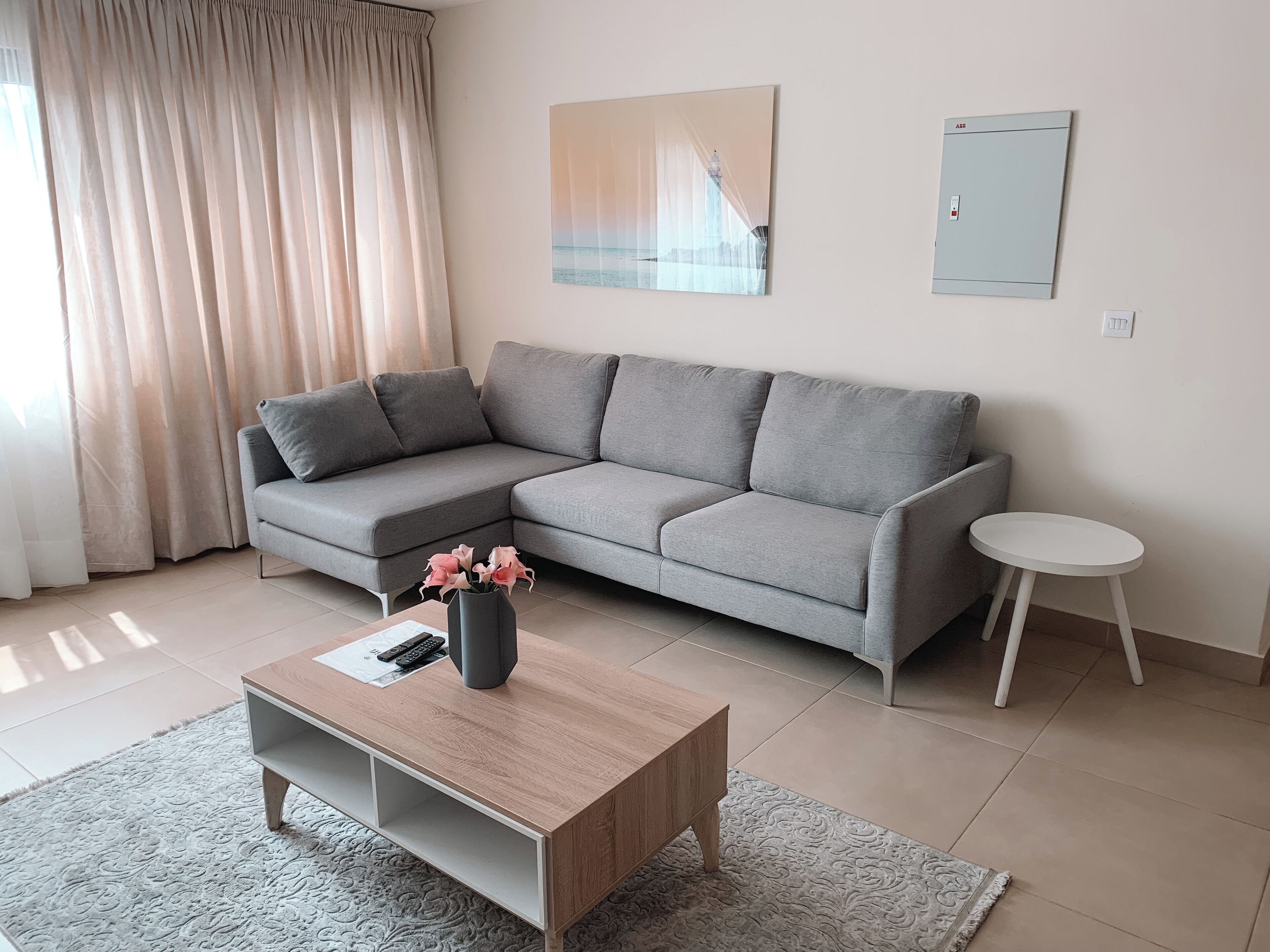 Ein modernes Wohnzimmer mit einem grauen Ecksofa, einem Holztisch, einem kleinen weißen Beistelltisch und einer Vase mit rosa Blumen.