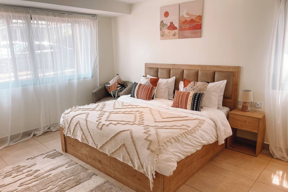 Une chambre à coucher avec un lit en bois, de nombreux oreillers, une table de chevet avec une lampe, et une vue sur l’extérieur à travers des rideaux légers.
