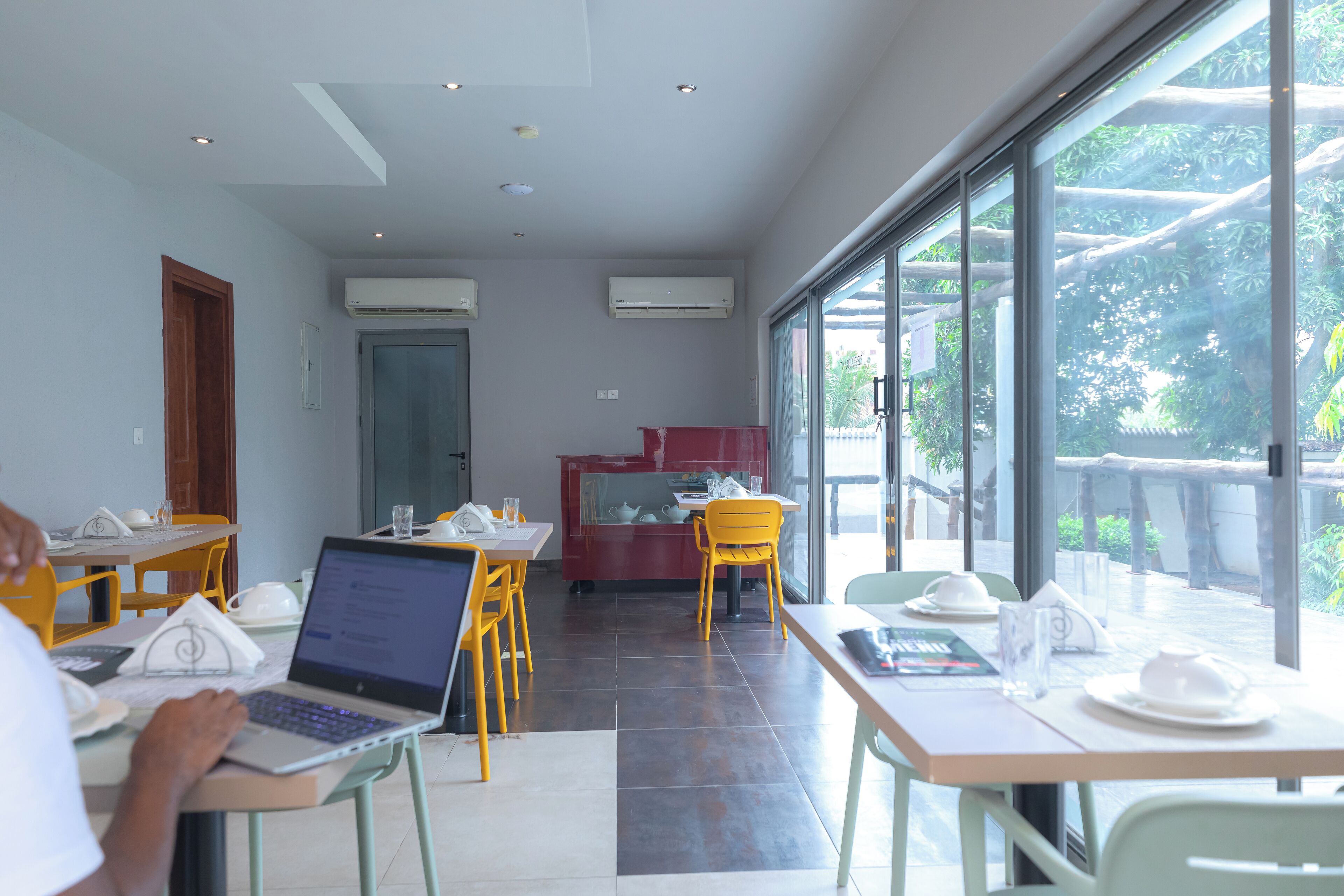 Un restaurante con mesas y sillas, una computadora portátil sobre una mesa, y vistas a árboles a través de amplias ventanas.