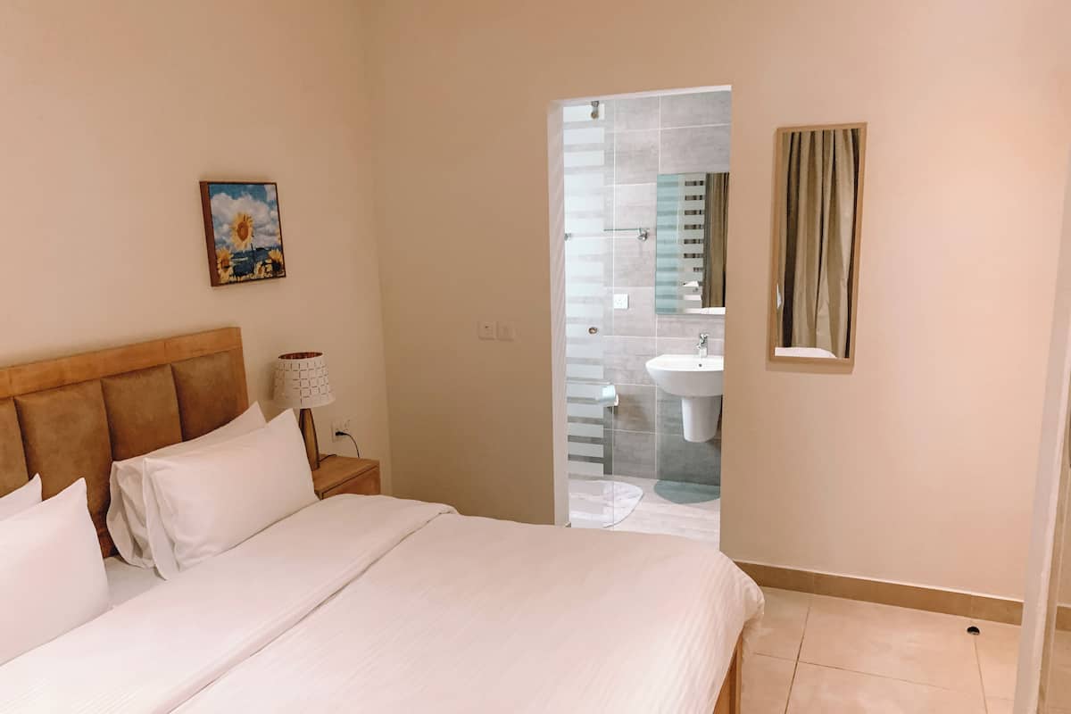 Une chambre à coucher avec un lit, une table de chevet, une lampe, un tableau, et une salle de bain visible à travers une porte ouverte.