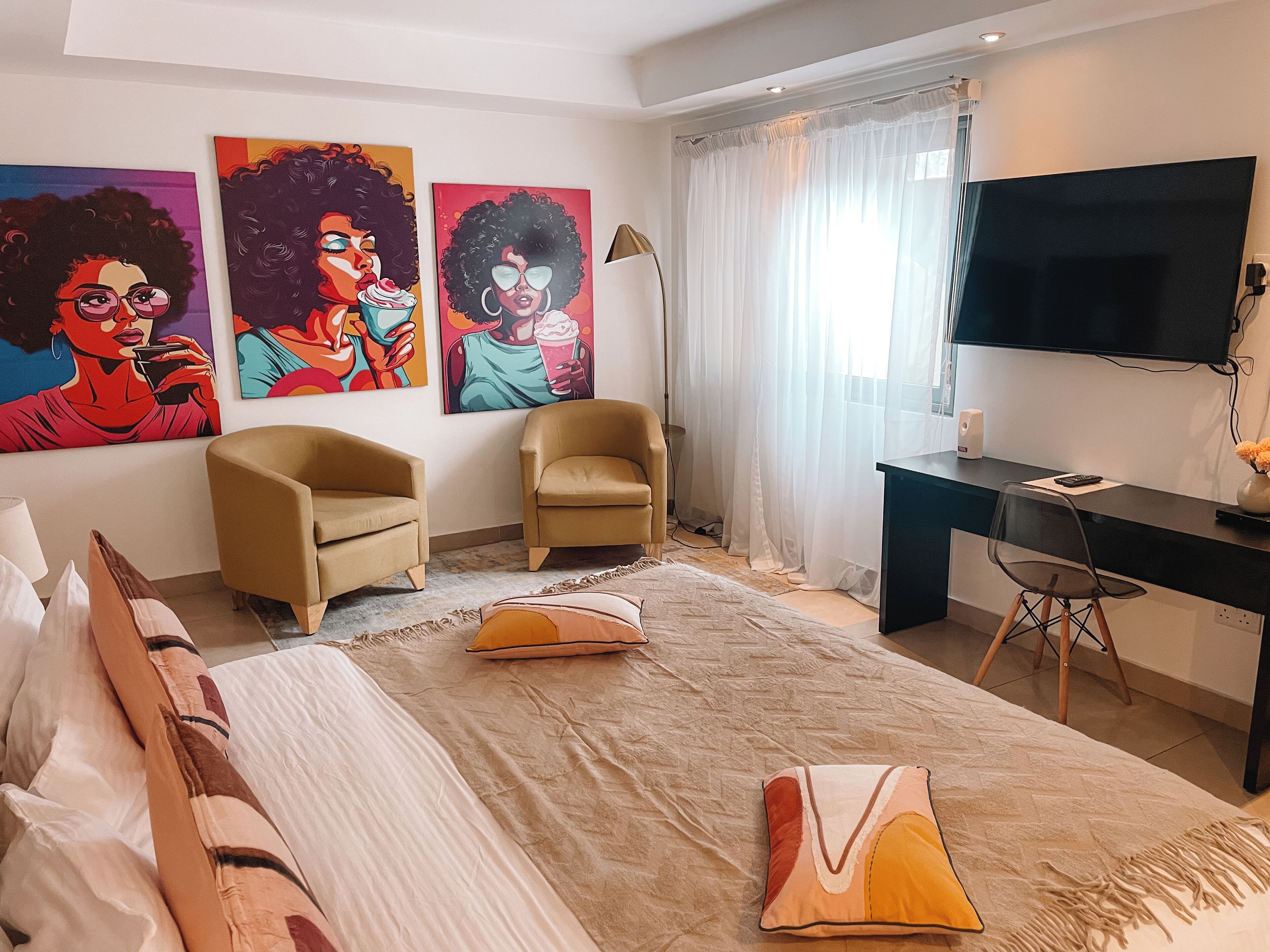 A hotel room with a bed, two armchairs, a desk with a computer, a TV, and three colorful paintings on the wall.