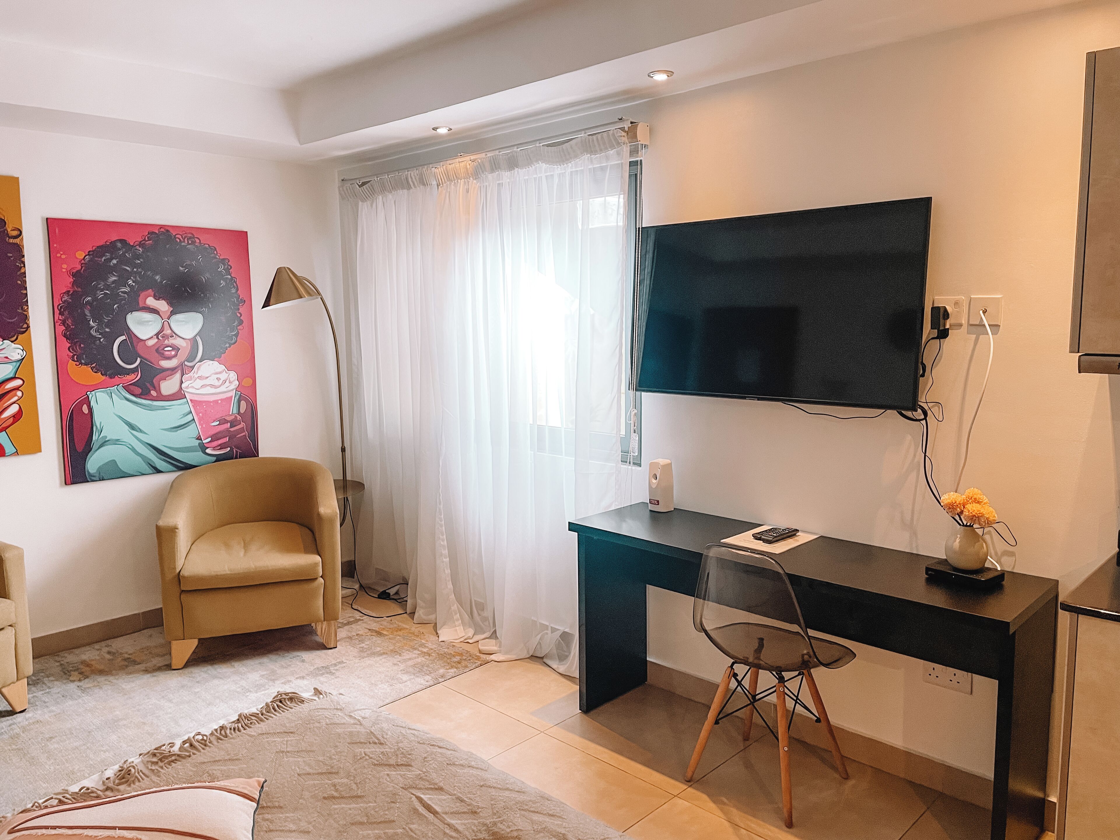 Una sala moderna con un sillón beige, un escritorio con silla, un televisor de pantalla plana y un ventanal con cortinas translúcidas.
