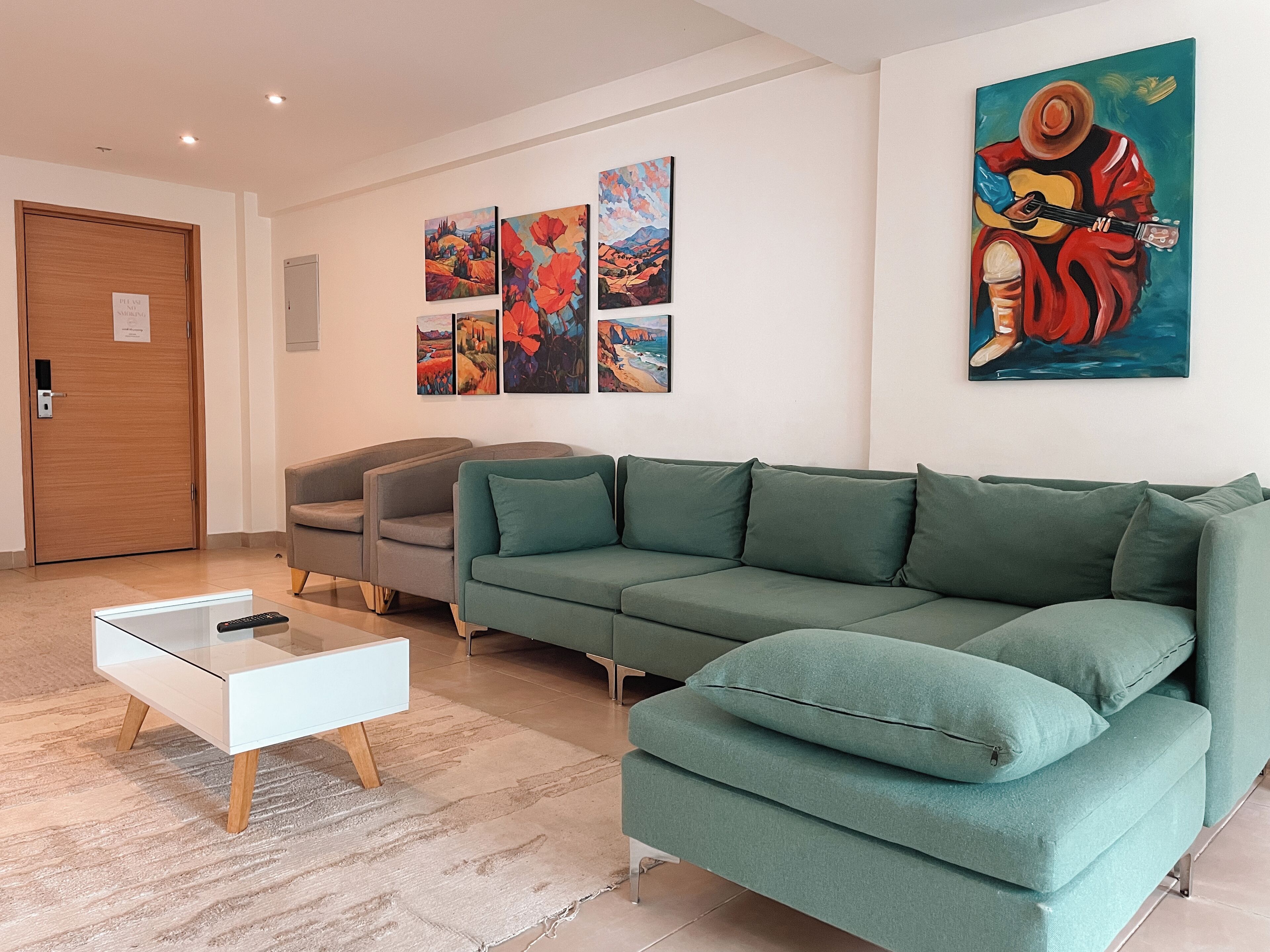 A modern living room with a green sofa, a white side table, and a remote control on it. There are three paintings on the wall and a large painting of a person playing a guitar.