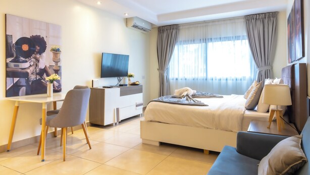 A modern hotel room with a bed, a desk, a chair, a television, and a large window with curtains. - Sky Suites Airport (Accra)