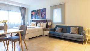 A modern hotel room with a bed, a sofa, a dining table, and chairs. There are three paintings on the wall above the bed. - Sky Suites Airport (Accra)