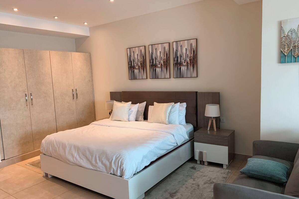 Une chambre moderne avec un grand lit, une armoire, une table de chevet, une chaise et deux œuvres d’art accrochées au mur.