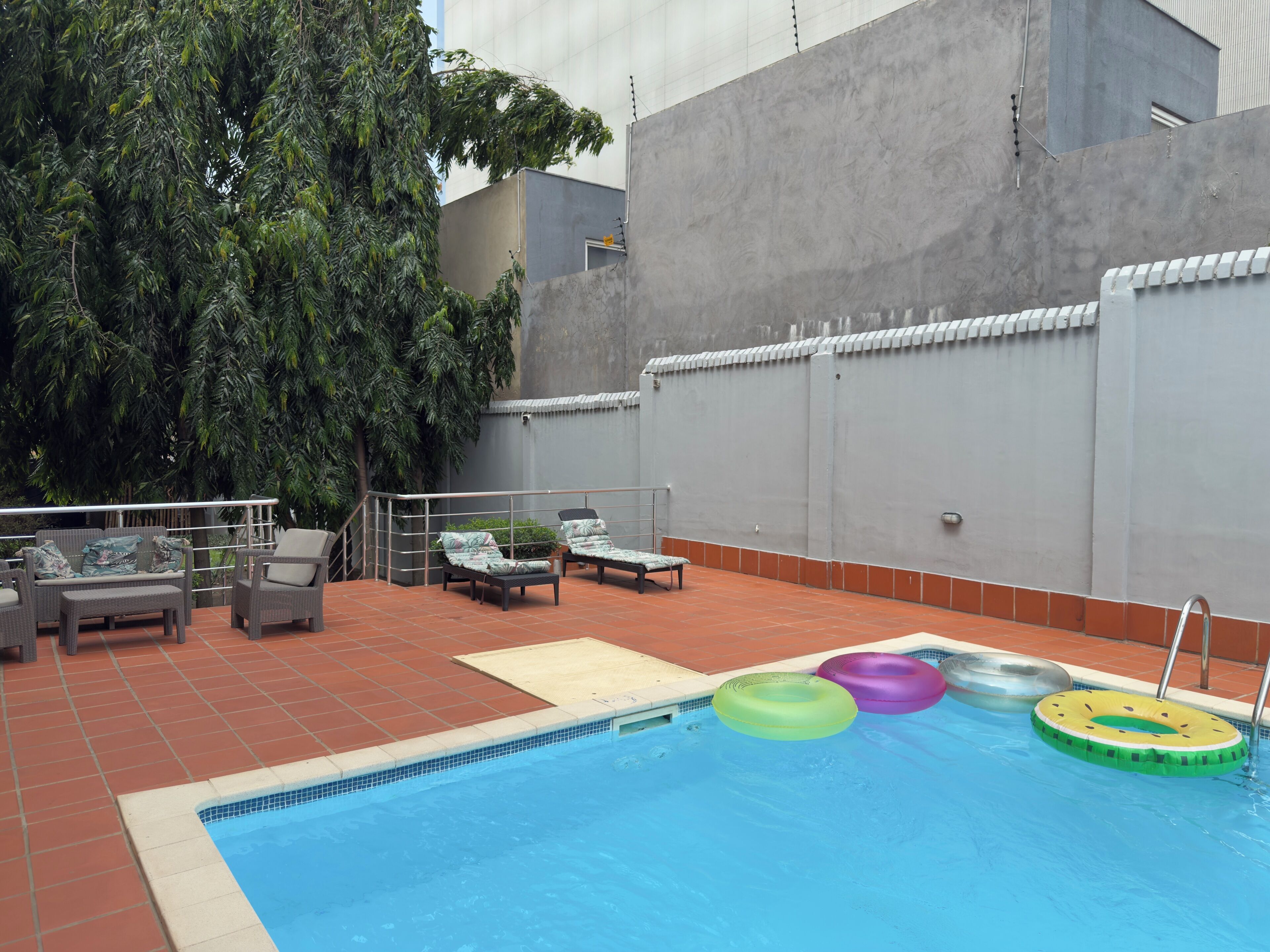 Una piscina con sillas de descanso y flotadores, rodeada por un patio empedrado y un muro gris.