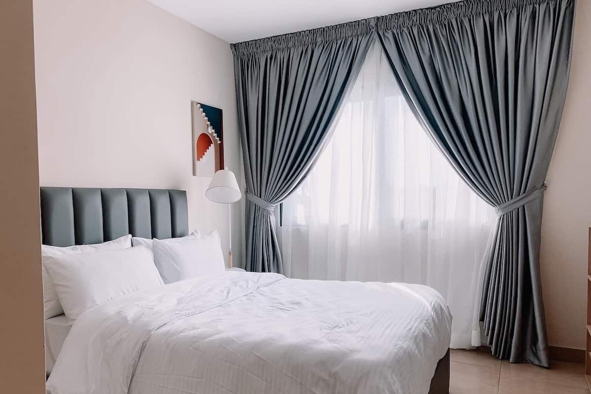 Un lit bien fait, avec du linge de lit blanc, une tête de lit rembourrée de couleur grise et une lampe fixée au mur.