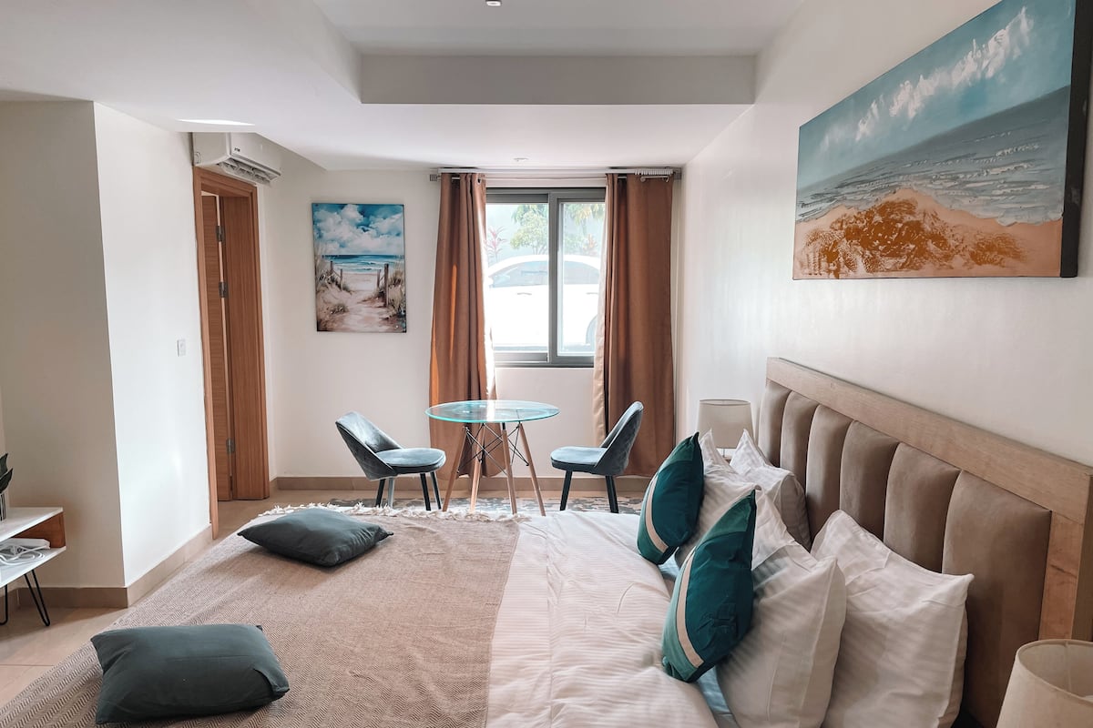 Une chambre d’hôtel avec un grand lit, une petite table avec des chaises, un tableau au mur et une fenêtre avec des rideaux.