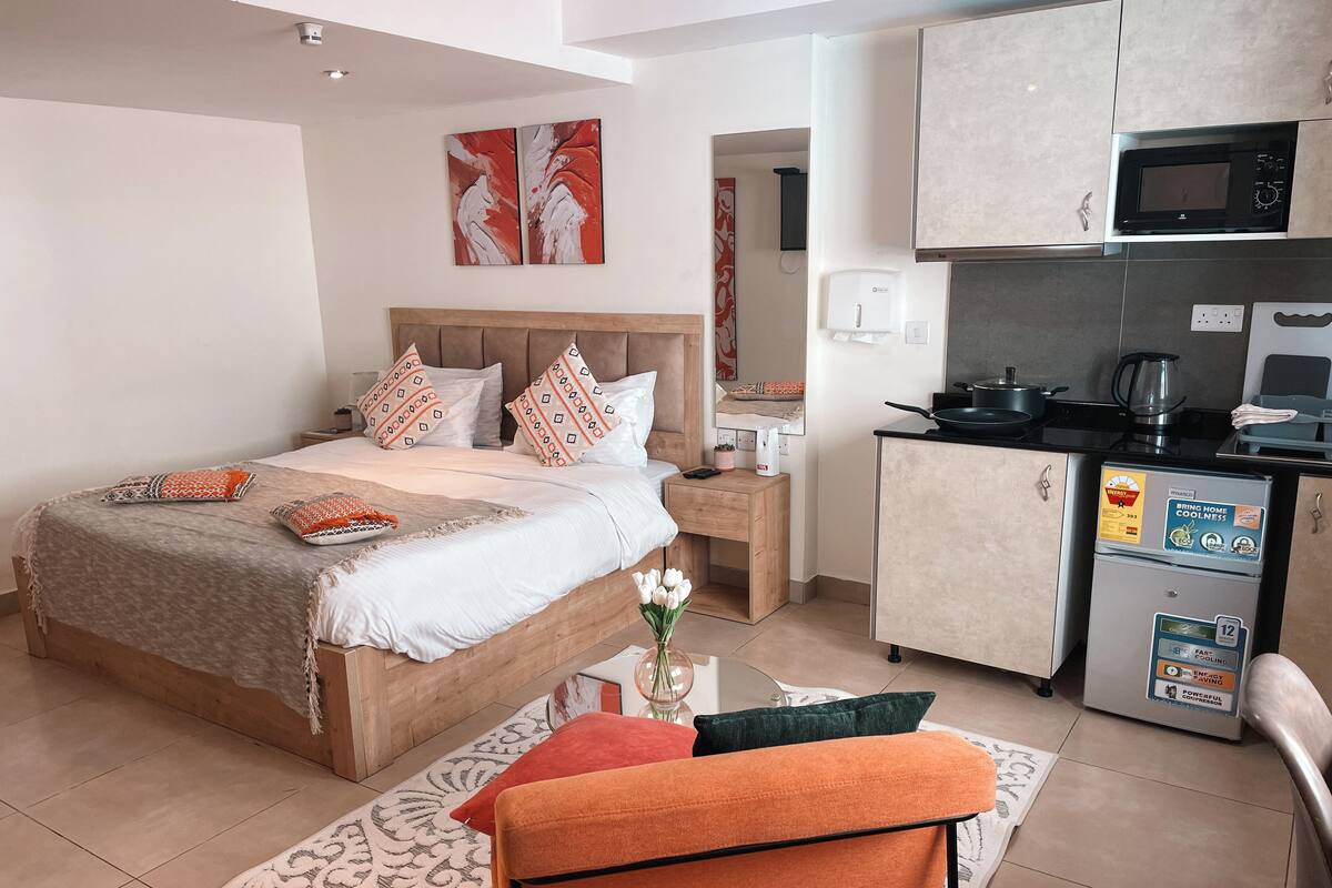 Une chambre à coucher comprenant un lit, une table de chevet et une petite table sur laquelle est posé un vase de fleurs. À côté se trouve une kitchenette équipée d’un four à micro-ondes, d’une cuisinière et d’un réfrigérateur.