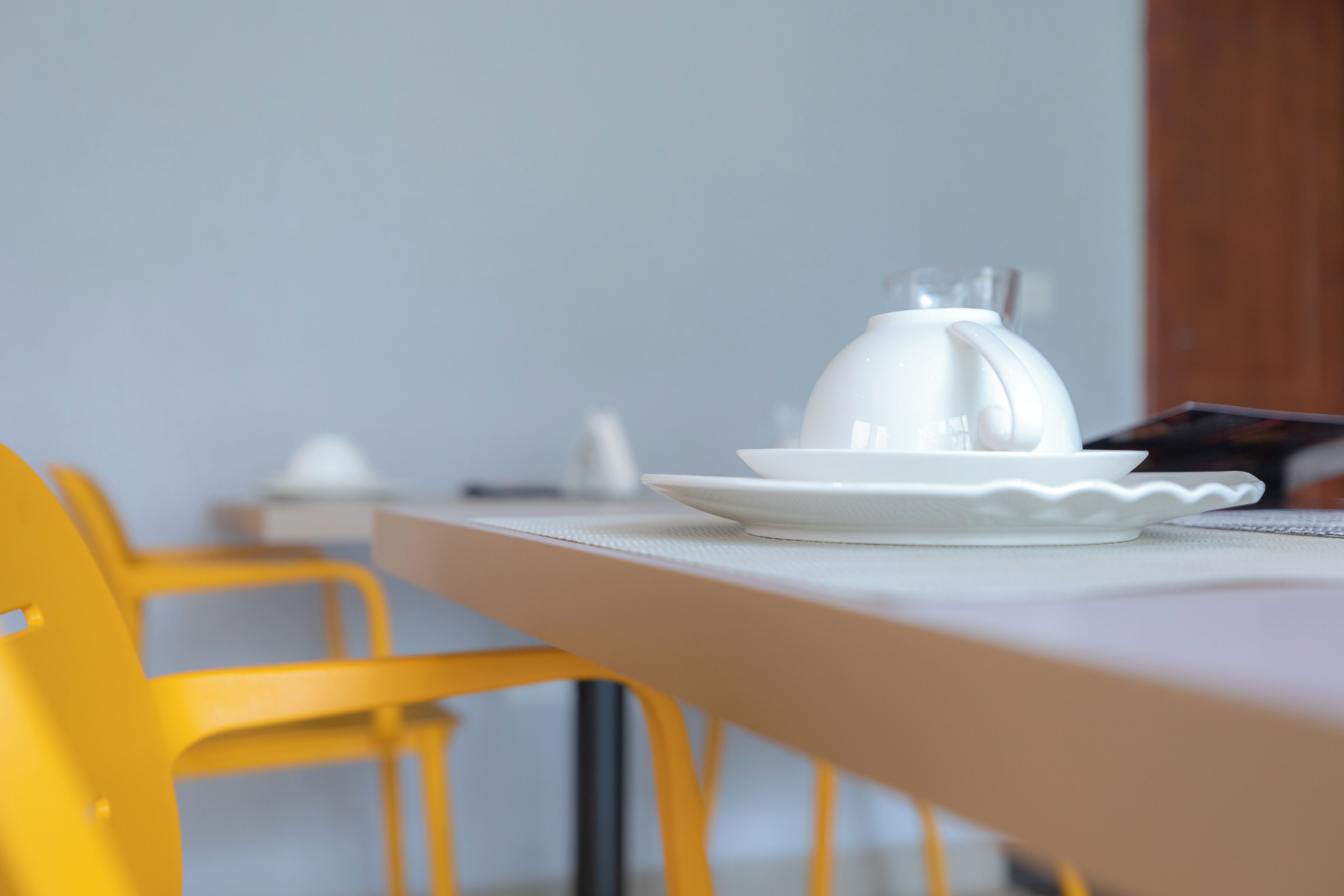 A dining table with a white pitcher and plates, a yellow chair, and a wooden cabinet in the background.
