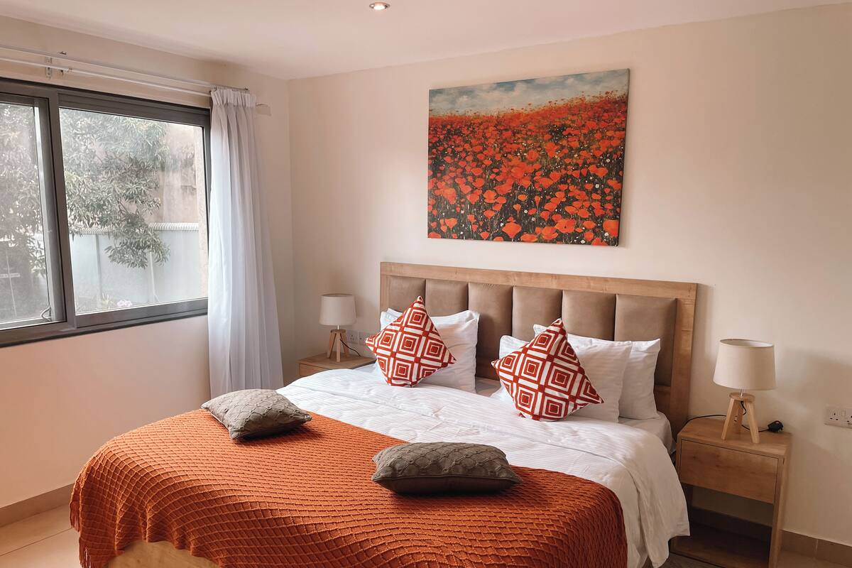 Un lit bien fait, recouvert d’une couverture à motifs, un tableau accroché au-dessus et une fenêtre avec des rideaux.