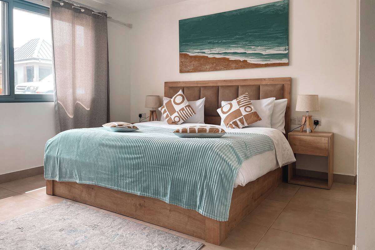 Un lit bien fait, recouvert d’une couverture à rayures, une tête de lit ornée d’un tableau représentant la mer, et une table de chevet avec une lampe.