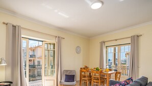 Un salon comprenant une table à manger, un canapé et un fauteuil. Il y a un balcon d’où l’on peut admirer la vue sur les immeubles.