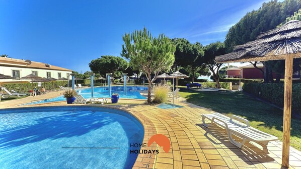 A swimming pool area with lounge chairs and umbrellas, surrounded by greenery and a building.