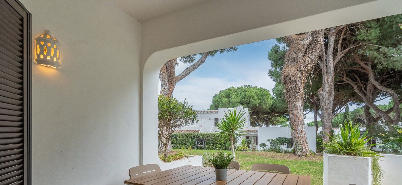 Un patio con una mesa y sillas de madera, una planta en maceta y una lámpara de pared.