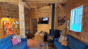 Interior acollidor d'una cabana amb dos sofàs blaus, una taula de fusta, una llar de foc i una televisió penjada a la paret.