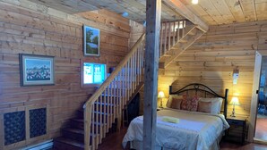 Dormitori d'una cabana de fusta amb un llit, una tauleta de nit, una làmpada, una escala de fusta i una finestra amb vista a arbres.