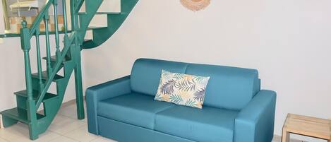 A teal-colored sofa with a leaf-patterned pillow in a room with a teal staircase and a wooden side table.