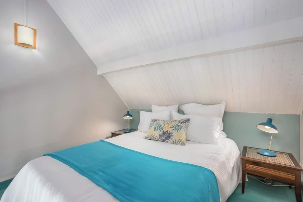 Ein Schlafzimmer mit schrägen Wänden, einem Bett mit weißer Bettwäsche und einer blauen Decke, zwei Nachttischen mit Lampen sowie einer Wandleuchte.