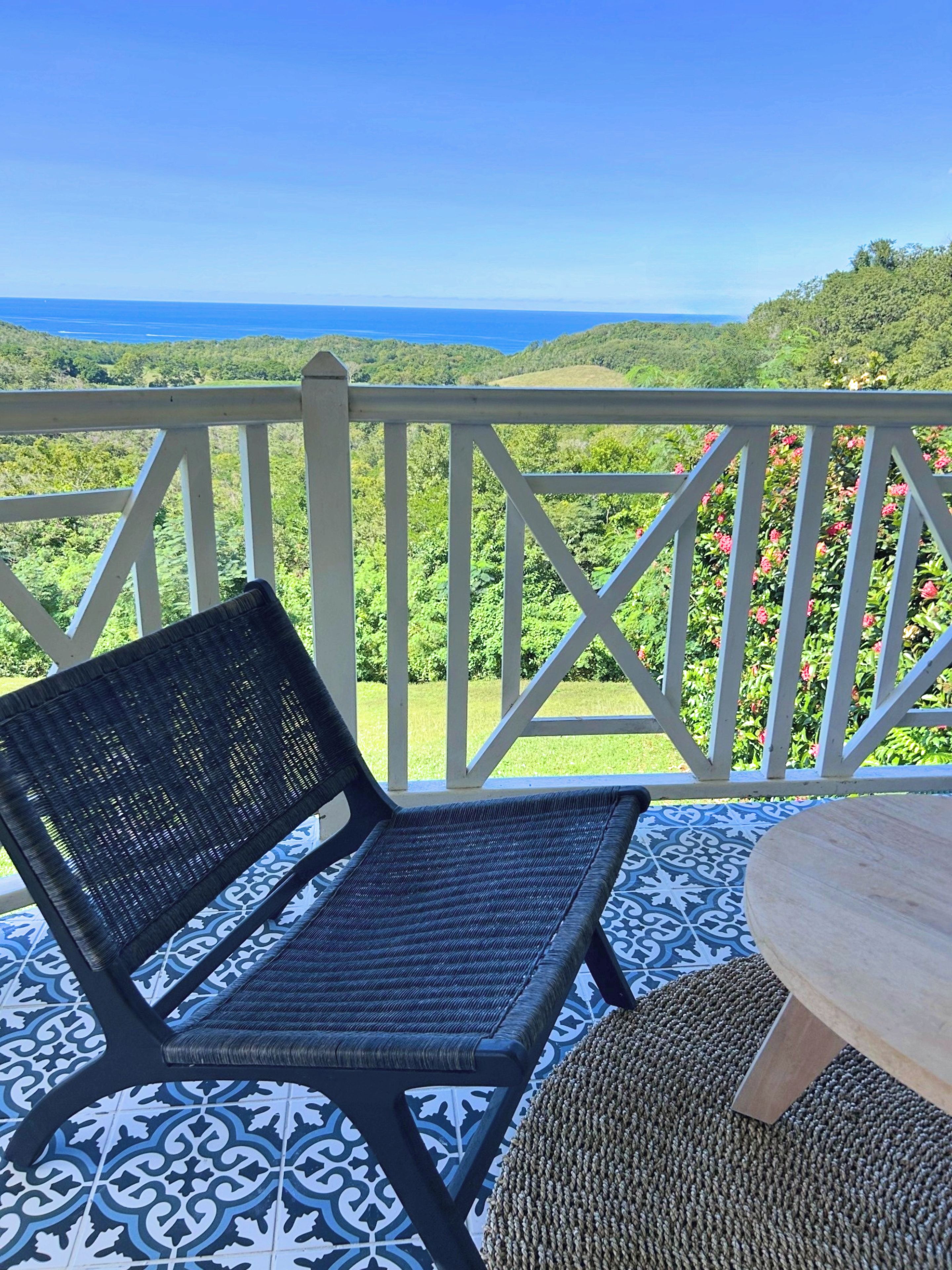 A balcony with a wicker chair and a small table, overlooking a lush landscape and the ocean.